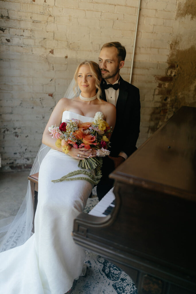 Bride and groom sit at an antique piano with vibrant flowers.